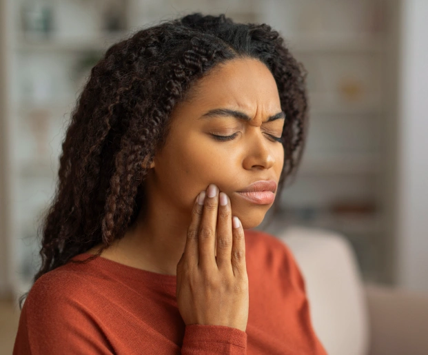 Woman in red shirt touching her cheek, appearing to experience tooth or jaw pain indoors.